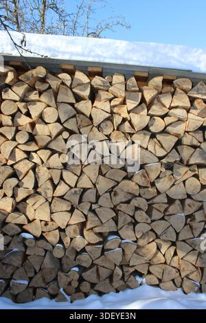 Ein großer Stapel ordentlich angeordneten Brennholzes steht in ländlicher Umgebung. Das Holz wird in gleichmäßige Stücke geschnitten und gegen ein Gebäude gestellt. Schnee ist sichtbar Stockfoto
