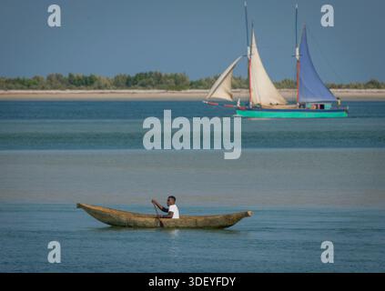 Ein madagassischer Mann in einem Kanu navigiert durch das flache Wasser des Mosambik-Kanals vor Belo Sur Mer. Direkt dahinter bewegt sich ein größeres traditionelles Fischerboot mit Segeln, bekannt als Lancha oder Botsy, vor der Küste. Beide Schiffe sind für die lokalen Fischereigemeinden von Vezo von entscheidender Bedeutung; das kleinere Kanu wird für die Fischerei und den Transport in Lagunen genutzt, während die größeren Segelboote schwere Lasten wie Salz oder landwirtschaftliche Erzeugnisse transportieren können und Küstengemeinden miteinander verbinden, die nicht über die Straße erreichbar sind. Stockfoto