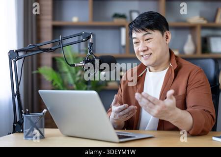 Asiatischer Mann, der ansprechende Inhalte in seinem Heimstudio erstellt, in ein professionelles Mikrofon spricht und einen Laptop ansieht, während er sendet oder rekordiert Stockfoto