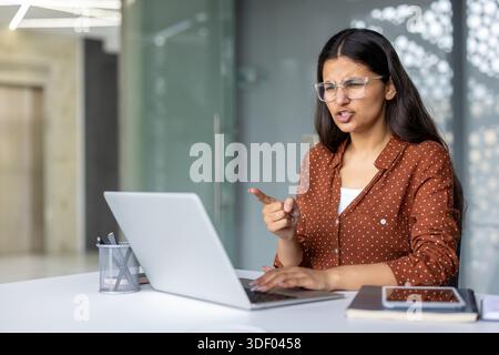 Junge indianerin, die Wut und Frustration während eines virtuellen Meetings ausdrückt, sich mit jemandem im Internet streitet und nicht einverstanden erklärt, während sie von einem aus aus arbeitet Stockfoto
