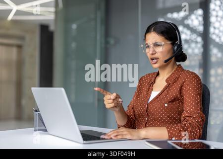 Junge Geschäftsfrau mit Headset und Brille, die in einem modernen Büro wütend auf einen Laptop zeigt und dabei Stress und Frustration zeigt Stockfoto