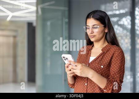 Eine junge Frau mit Brille und einem Polka Dot-Shirt tippt auf ihrem Handy, während sie in einem modernen Büro steht und sich mit der Kommunikation beschäftigt Stockfoto