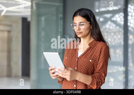 Eine junge professionelle Frau in Brille und einem Polka-Dot-Shirt steht in einem modernen Büro und konzentriert sich auf ein digitales Tablet, während sie arbeitet, surft und verbindet Stockfoto