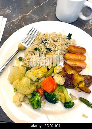 Teller mit gemischten Speisen mit gebratenem Reis, gebratenen Kartoffeln, Mini-Würstchen und verschiedenen gedämpften Gemüsesorten auf einem Esstisch Stockfoto