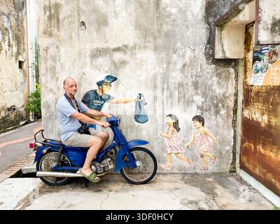 Männlicher Tourist auf einem Vintage-blauen Motorrad posiert vor einem interaktiven Wandgemälde mit Kindern und Essensreiter Concubine Lane, Ipoh malaysia Stockfoto
