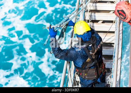 Seeleute, die die Pilotenleiter an der Seite eines Frachtschiffes nach dem Ausstieg des Piloten auf See sichern, Blick von oben, raues blaues Meer im Hintergrund. Stockfoto