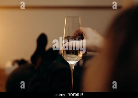 Eine Hand, die ein Flötenglas mit Sekt oder Champagner hält. Das Bild konzentriert sich auf das Getränk und die Glaswaren vor einem warm beleuchteten, verschwommenen Inter Stockfoto