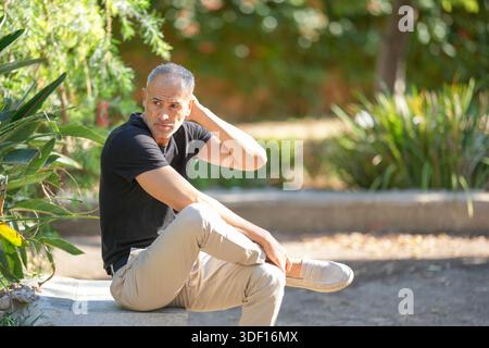 Ein Mann mit schwarzem Hemd und Khaki-Hose sitzt auf einer Steinbank im Park. Er ist in Gedanken verloren, mit der Hand auf dem Kopf. Der Park ist sehr schön Stockfoto