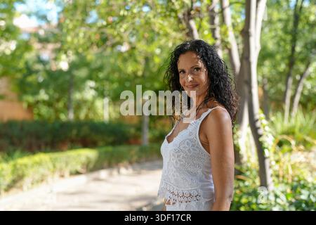 Eine Frau mit lockigen Haaren steht in einem Park und trägt ein weißes Tanktop. Sie lächelt und sieht in die Kamera Stockfoto