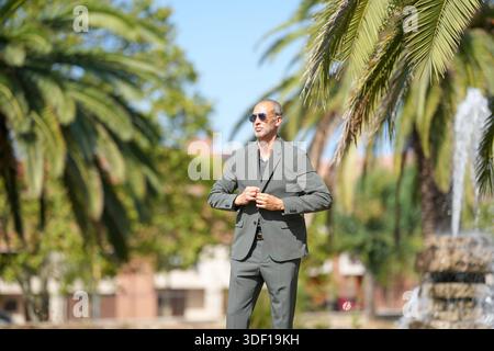 Ein Mann im Anzug steht vor einem Brunnen. Er trägt eine Sonnenbrille und passt seine Jacke an. Die Szene spielt in einem tropischen Ort, mit Stockfoto