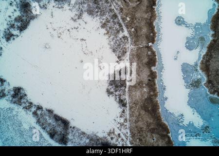 Die Luft der Winterlandschaft fängt den Übergang zwischen einem unberührten schneebedeckten Feld und einem gefrorenen blauen See ein, der durch einen rostigen Gürtel aus trockenem Schilf getrennt wird. Die Komposition bietet ausreichend Platz für saisonale Nachrichten oder Umgebungstexte. Stockfoto