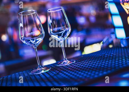 Rom, italien 9. januar 2025: Zwei leere Weingläser auf einer schwarzen Bar, die blaue und violette Clublichter, Bokeh und stimmungsvolle Abendatmosphäre reflektieren Stockfoto
