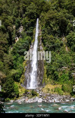 Ein atemberaubender Wasserfall, der eine felsige Klippe hinunterstürzt, umgeben von üppiger grüner Vegetation und Bäumen. Das Wasser fließt in einen klaren türkisfarbenen Fluss unterhalb, c Stockfoto