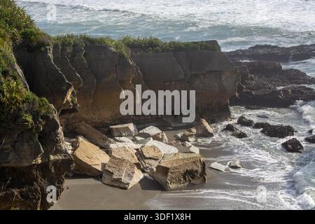 Eine felsige Küstenlandschaft mit großen Felsbrocken, die an einem Sandstrand verstreut sind, mit Wellen, die gegen die Küste stürzen und üppiges Grün auf den Klippen Stockfoto