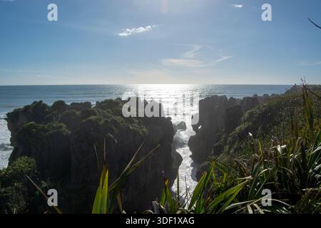 Ein malerischer Blick auf die Küste mit zerklüfteten Klippen und üppigem Grün im Vordergrund, wobei die Sonne hell über dem Meer scheint, was ein Schimmer erzeugt Stockfoto
