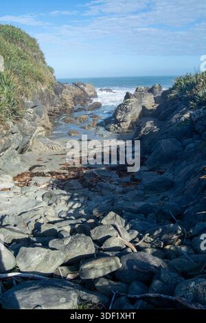 Eine felsige Küstenlandschaft mit einem schmalen Pfad, der zum Meer führt. Die Szene zeigt große Felsbrocken, Sandflächen und Algen, die am Ufer verstreut sind. Stockfoto