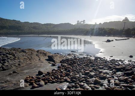 Eine ruhige Strandszene mit glatten Felsen und Kieselsteinen entlang der Küste, mit sanften Wellen, die auf den Sand plätschern. Üppige grüne Hügel erheben sich im Hinterland Stockfoto