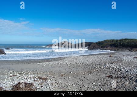 Eine ruhige Strandszene mit sanften Wellen am Ufer, Kieselsteinen über den Sand und felsigen Klippen im Hintergrund unter einem klaren Wasser Stockfoto