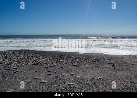 Eine ruhige Strandszene mit einem felsigen Ufer mit glatten Kieselsteinen, sanften Wellen am Wasserrand und einem klaren blauen Himmel über dem Wasser. Stockfoto