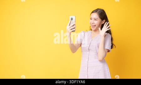 Fröhliche asiatische Frau nimmt Selfie und winkt Hand während Videoanruf mit Smartphone im Studio, lächelnde junge Frau auf hellgelbem Hintergrund, Onli Stockfoto