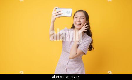 Fröhliche asiatische Frau nimmt Selfie und winkt Hand während Videoanruf mit Smartphone im Studio, lächelnde junge Frau auf hellgelbem Hintergrund, Onli Stockfoto