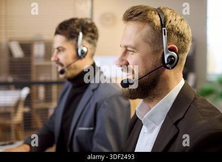 Lächelnde männliche Supportmitarbeiter mit Headsets, die in modernen Callcentern im Büro arbeiten Stockfoto