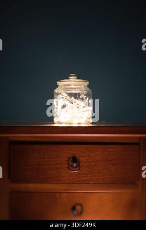 Ein minimalistisches Stillleben mit einem transparenten Glasgefäß, gefüllt mit warmen, leuchtenden Lichtern, auf einer Holzschublade vor dunklem Hintergrund. Stockfoto