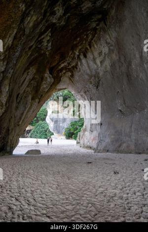 Eine malerische Aussicht aus dem Inneren einer großen Höhle mit Blick auf einen Sandstrand und üppiges Grün. Zwei Leute laufen am Strand, eingerahmt von den Höhlen Stockfoto
