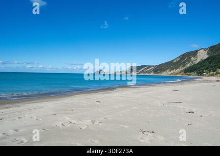 Eine ruhige Strandszene mit weichem Sand, sanften Wellen und einem klaren blauen Himmel. Die Küste ist von Hügeln gesäumt, was eine malerische Landschaft schafft Stockfoto