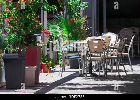 Auf der sonnendurchfluteten Terrasse, umgeben von üppigem Grün und leuchtend rosa Blumen, stehen Café-Stühle und Tische aus Metall. Die friedliche Umgebung lässt eine ruhige Mutter vermuten Stockfoto