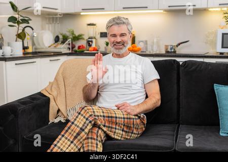 Ein Mann mittleren Alters zu Hause winkt zur Kamera, sagt Hallo und beginnt mit einem freundlichen Lächeln Online-Kontakt. Reifer Mann auf dem Sofa schaut auf die Linse nickt spricht kurze Begrüßung warme Stimmung offener Ton ruhiger Atem ruhig. Stockfoto