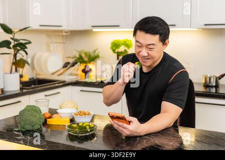 Asiatischer Mann in der Küche isst Brokkoli-Snack und überprüft Gesundheitsnachrichten auf orange Smartphone. Ein erwachsener Mann mittleren Alters genießt kalorienarme Snacks drinnen und fühlt sich motiviert, den Ernährungsplan und die Änderung des Lebensstils zu befolgen. Stockfoto