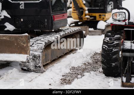 Details zu Baumaschinen, die in der kalten Jahreszeit auf verschneiten Oberflächen auf der Außenarbeitsstelle Stahlketten des Baggers und ein kompaktes Fahrzeugrad zeigen Stockfoto