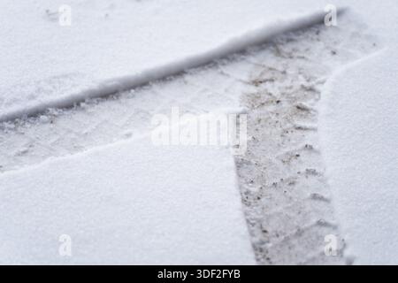 Schneebedeckter Boden mit sichtbaren Spuren von Fahrzeugreifen, die durch dünne Schneeschicht schneiden und Schmutzoberflächen freilegen, wodurch die Beschaffenheit des Winterbodens und die tran-Struktur entstehen Stockfoto