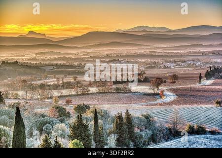 Eine ruhige Winterlandschaft mit Frost, die die sanften Felder bedeckt. Die aufgehende Sonne strahlt ein warmes Leuchten über ferne Berge. Stockfoto