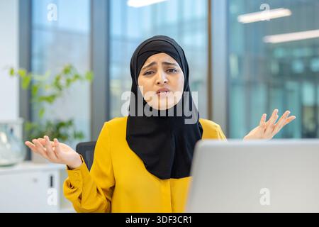Muslimische Geschäftsfrau mit Hijab und gelber Bluse, die mit einem verwirrten Gesichtsausdruck zuckte und sich in einem modernen Büro einen Laptop anschaut Stockfoto