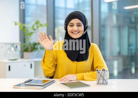 Glückliche muslimische Frau im Hijab, die ein Headset trägt und vor der Kamera winkt, am Schreibtisch sitzt und eine Videokonferenz abhält oder Kundensupport bietet Stockfoto