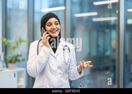 Junge muslimische Ärztin in einem weißen Mantel und Hijab lächelnd während eines Telefonats in einer hellen modernen Klinik, symbolisiert Telemedizin, professionelle CA Stockfoto