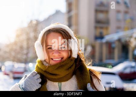 Junge Frau mit Ohrenschützern und warmem Schal, die draußen an einem sonnigen Wintertag steht. Stockfoto