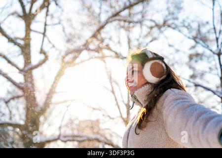 Junge lächelnde Frau in Kapselgehörschützern, die an einem hellen, schneebedeckten Tag im Winter den Lebensstil im Freien genießt Stockfoto