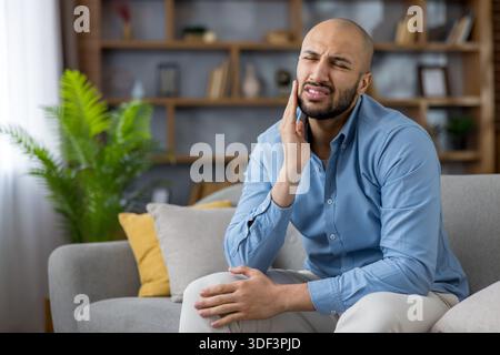 Junger Mann mit blauem Hemd und weißer Hose, der auf einem grauen Sofa in einem Wohnzimmer sitzt, grimmig und seine Wange berührt und einen intensiven Zahnschmerz erlebt Stockfoto