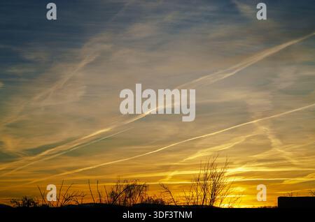 Dramatischer Sonnenuntergangshimmel voller Kondensstreifen und schimmernder Wolken, die goldene Farbtöne ausstrahlen Stockfoto