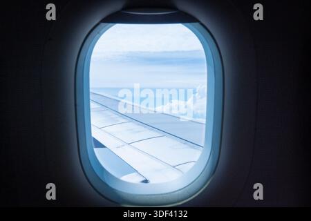 Ein Blick aus einem Flugzeugfenster, der den Flugzeugflügel und weiße Wolken vor einem blauen Himmel zeigt. Flug-, Tourismus- und Reisekonzept. Stockfoto