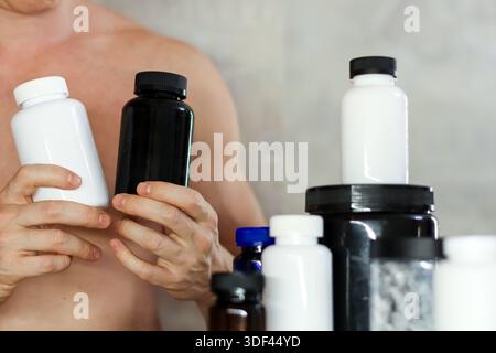 Mann, der weiße und schwarze Supplementflaschen vergleicht. Muskuläre männliche Haltung und Vergleich von weißen und schwarzen Kunststoff-Nahrungsergänzungsbehältern mit anderen in bac Stockfoto