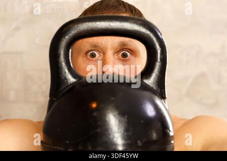 Schockierter Mann, der mit weiten Augen durch die Kettlebell starrt. Athletischer Mann mit überraschtem Ausdruck, der die Kettlebell geschlossen hält, Augen durch den Griff in Innenräumen sichtbar Stockfoto