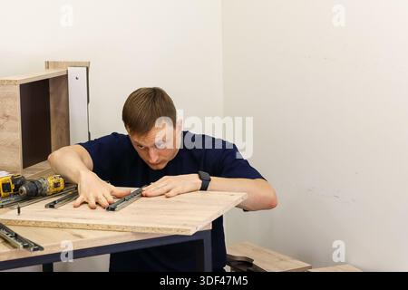 Mann, der die Führungen der Metallschublade an der Möbeltafel installiert. Ernsthafter Mann mit Smartwatch, der die Führungen der Metallschublade auf Holzmöbeln anbringt und ausrichtet Stockfoto