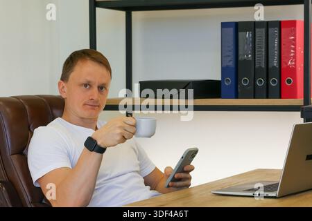 Ein lässiger gekleideter Mann schaut sich das Smartphone an und trinkt aus einer weißen Tasse am Schreibtisch mit Laptop. Stockfoto