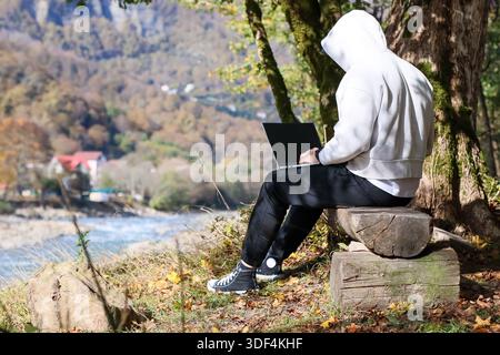 Rückansicht eines jungen Erwachsenen in Kapuzenkleidung, der auf einem Baum sitzt und auf einem Laptop mit Blick auf den Fluss und die Berge im Herbst tippt. Stockfoto