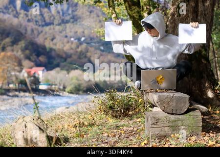 Die Person in weißem Hoodie und Sonnenbrille sitzt auf Holzholz an Baum und hebt zwei leere weiße Laken in jeder Hand mit Laptop auf Schoß im Herbst Stockfoto