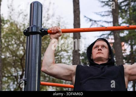 FIT männlich führt das Kinn auf der Pull-up-Leiste im Park mit Bäumen im Hintergrund aus Stockfoto